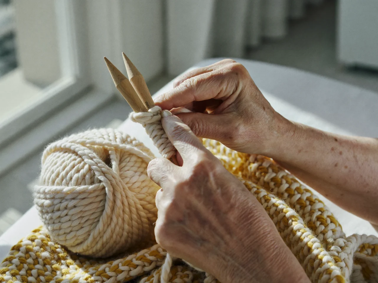 Mani anziane lavorano a maglia lana color panna e senape con ferri in legno.